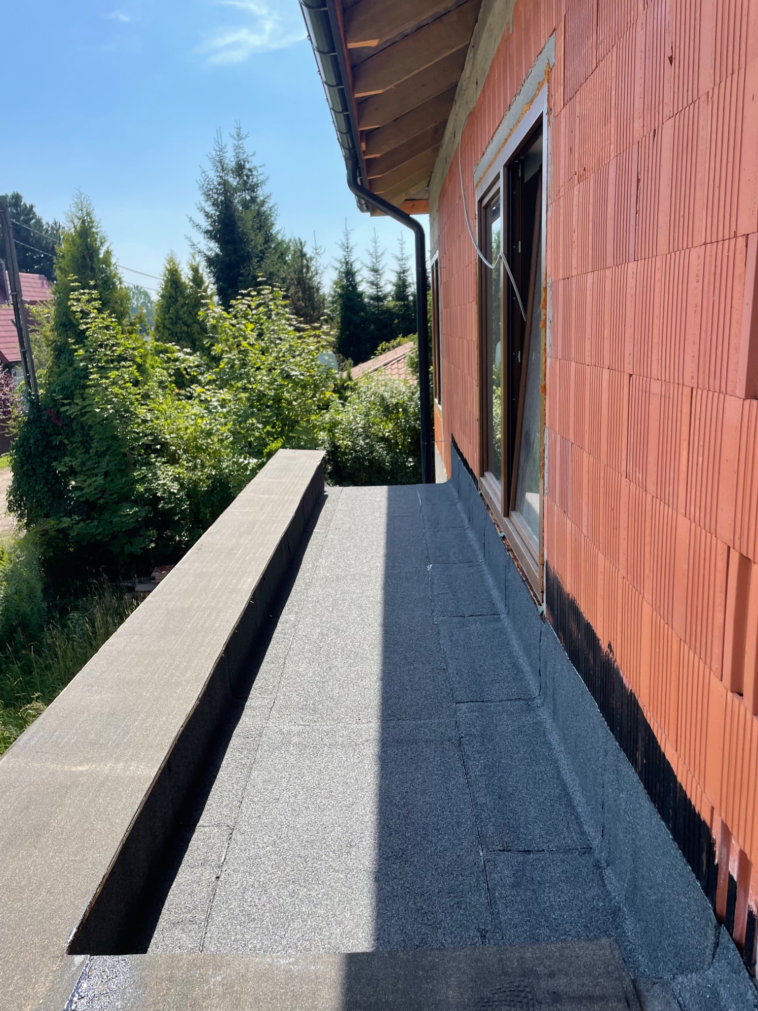 Wykończony taras z izolacją bitumiczną przy ścianie z pustaków ceramicznych z oknem, widok z góry, w tle zieleń i zabudowa jednorodzinna.