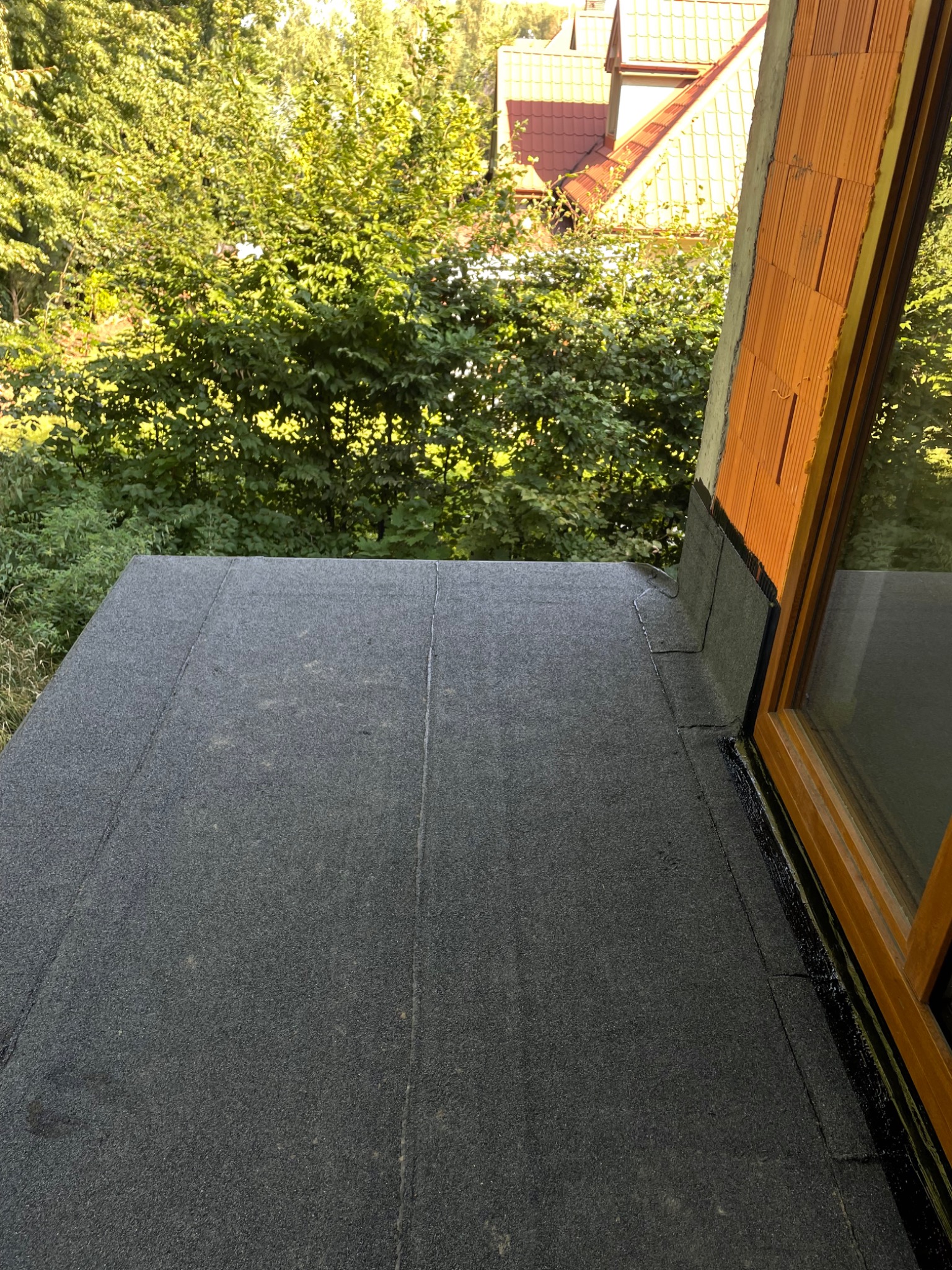 Wykończony płaski dach pokryty papą, z widocznym oknem dachowym i obróbkami blacharskimi, na tle zieleni.