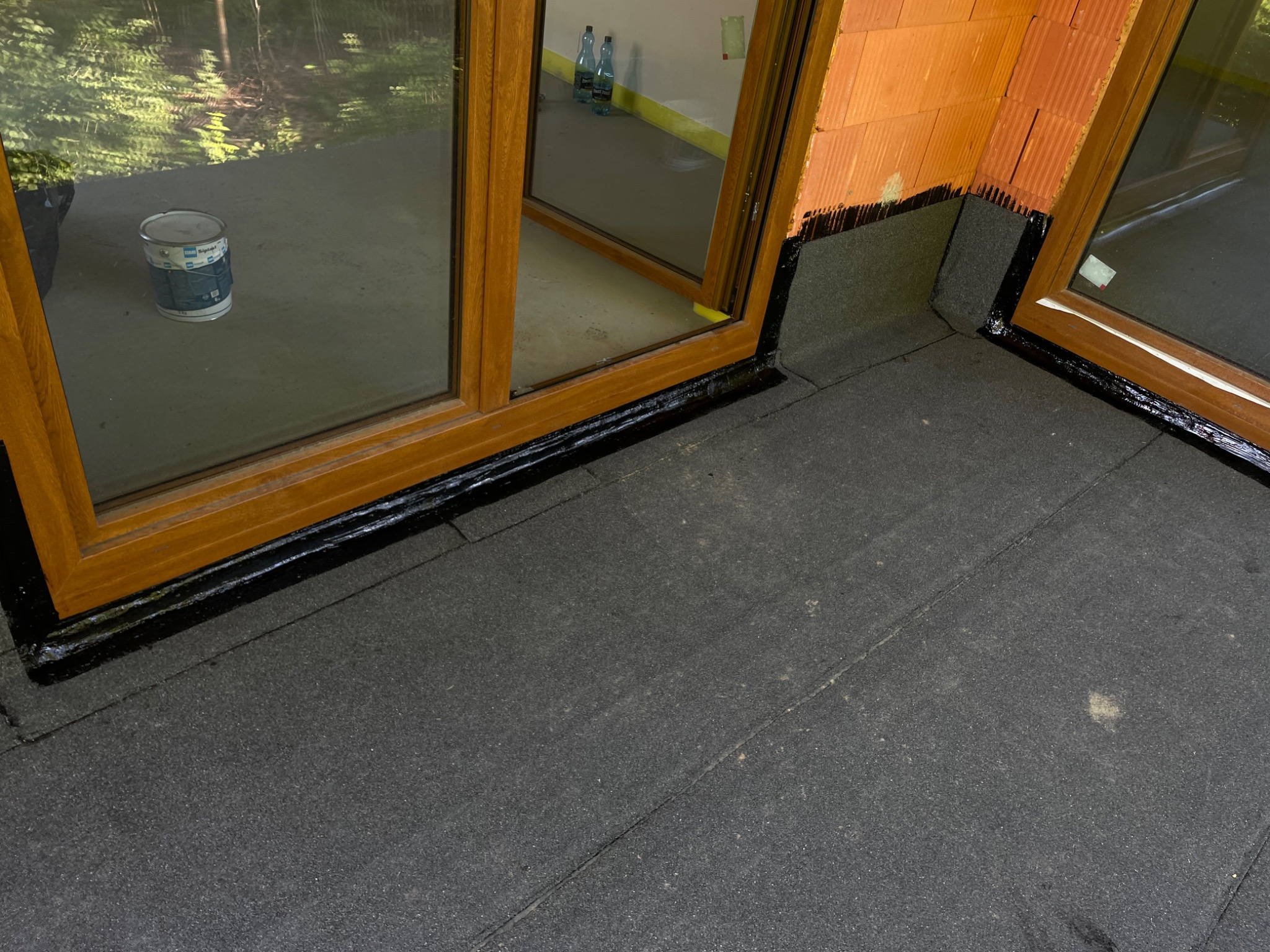 Uszczelnienie dachu z papy bitumicznej wokół okien dachowych, z widocznymi pomarańczowymi cegłami i odblaskami w szybach.