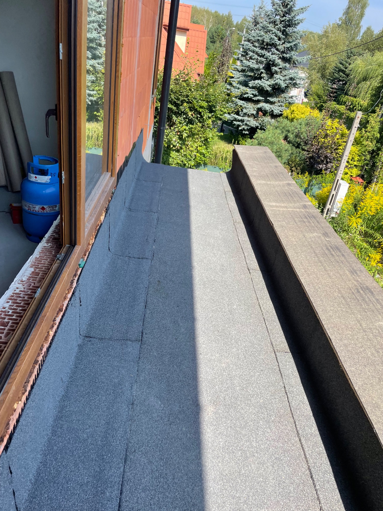 Płaski dach pokryty papą termozgrzewalną, widok z góry na łączenie z oknem dachowym, butla gazowa w tle.