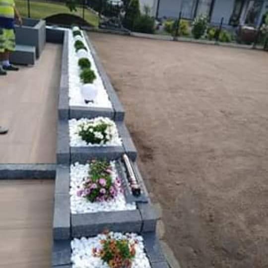 Kaskadowa konstrukcja z szarych betonowych bloków tworząca rabaty na białym żwirze, obsadzone różnokolorowymi kwiatami i kulistymi krzewami, z ziemią w tle.