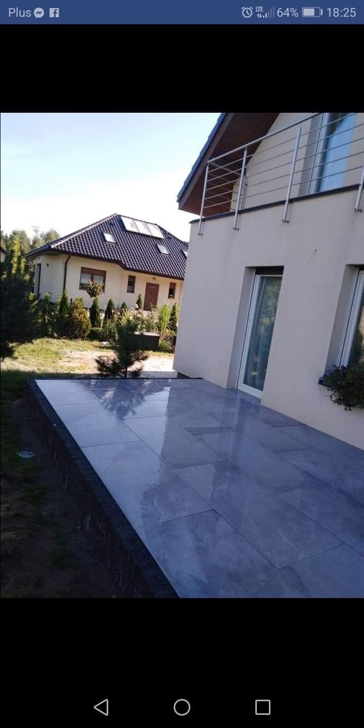 Wyłożony błyszczącymi, szarymi płytami taras przylegający do jasnego domu z balkonem, w tle drugi budynek.