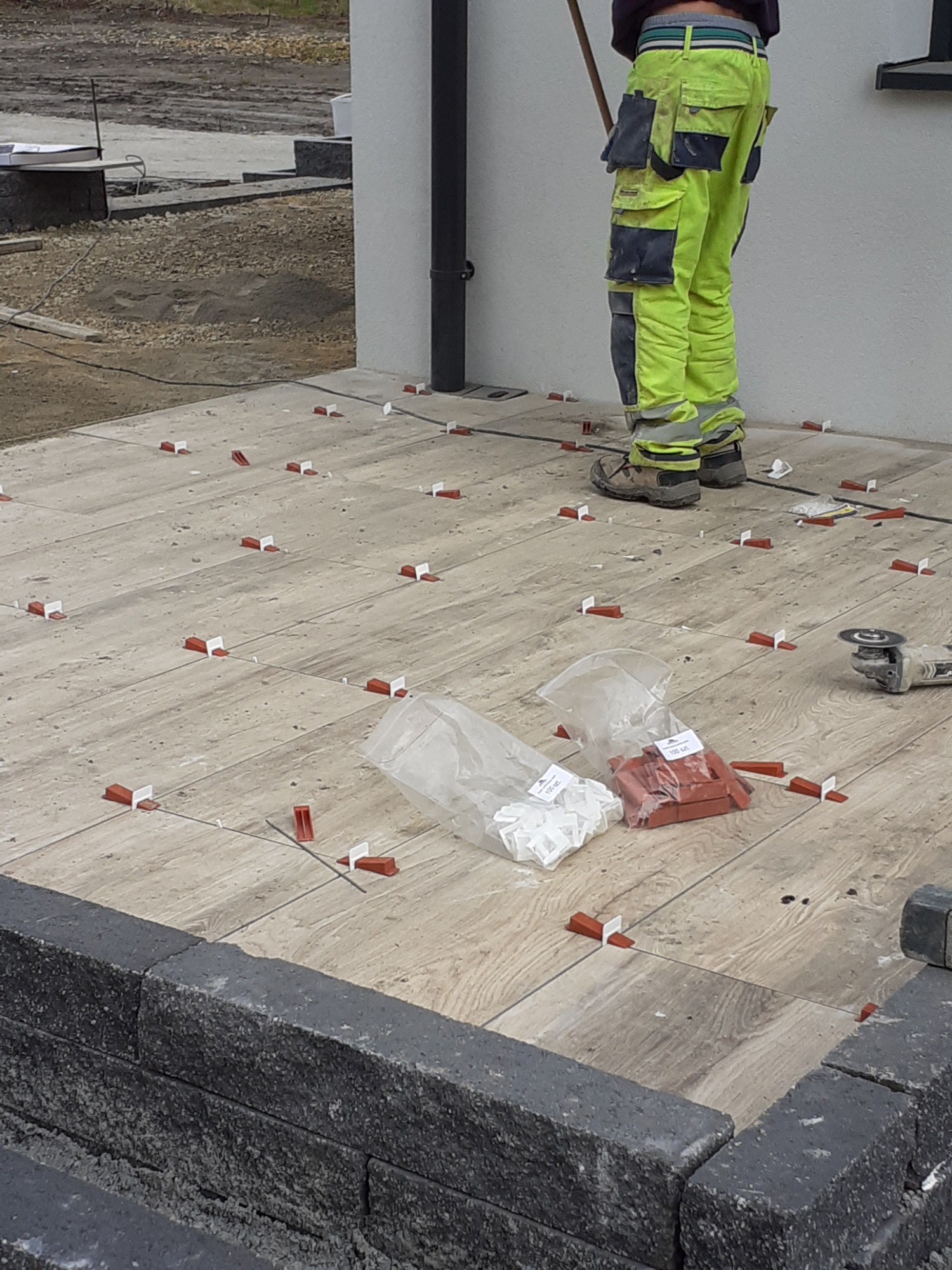 Układanie płytek na zewnątrz budynku, widoczne narzędzia i system poziomowania, pracownik w spodniach roboczych w kolorze neonowej zieleni.