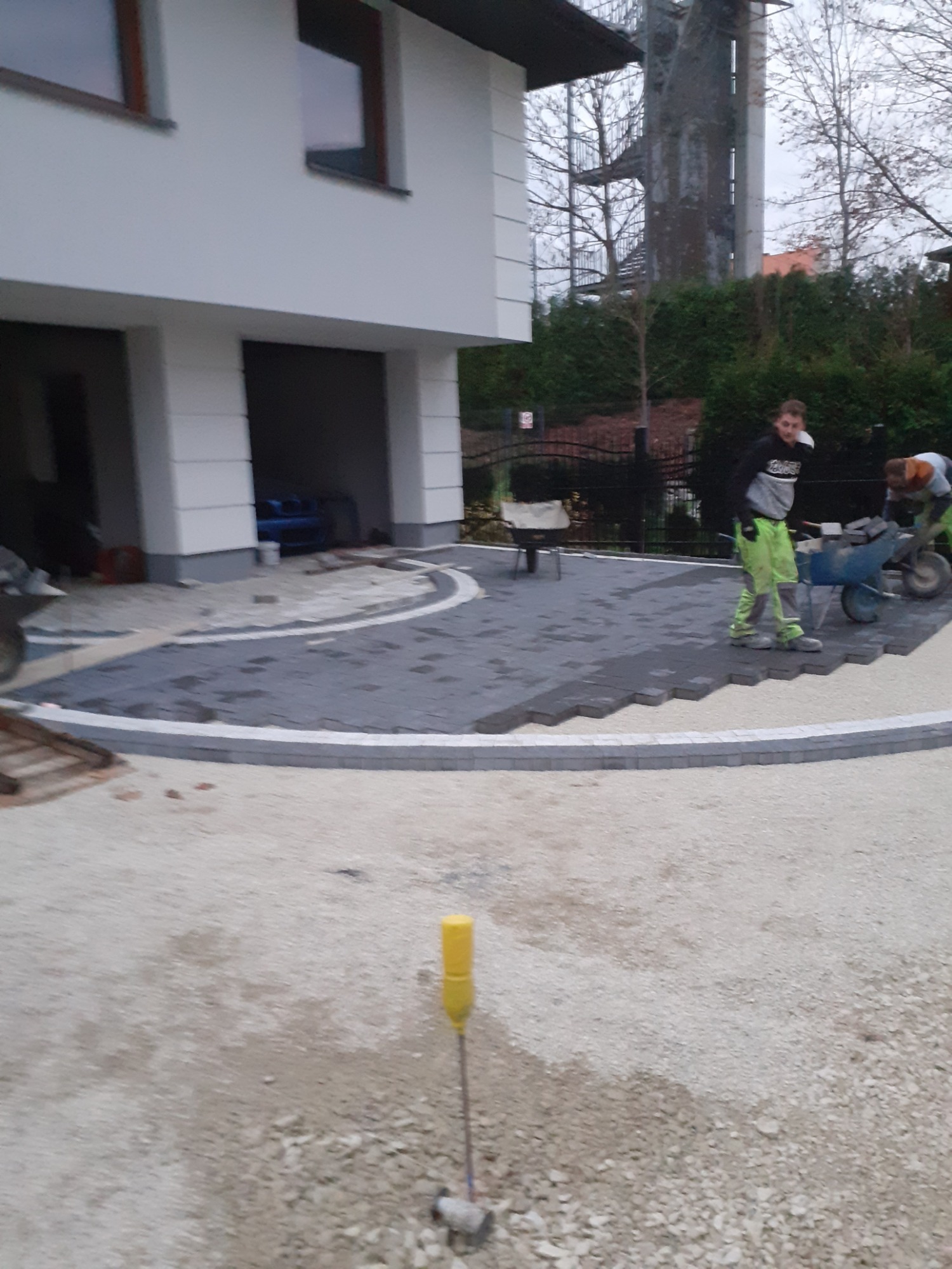 Układanie kostki brukowej na podjeździe przed domem, widoczni dwaj pracownicy z taczkami, w tle biały dom z garażem i wieża obserwacyjna.