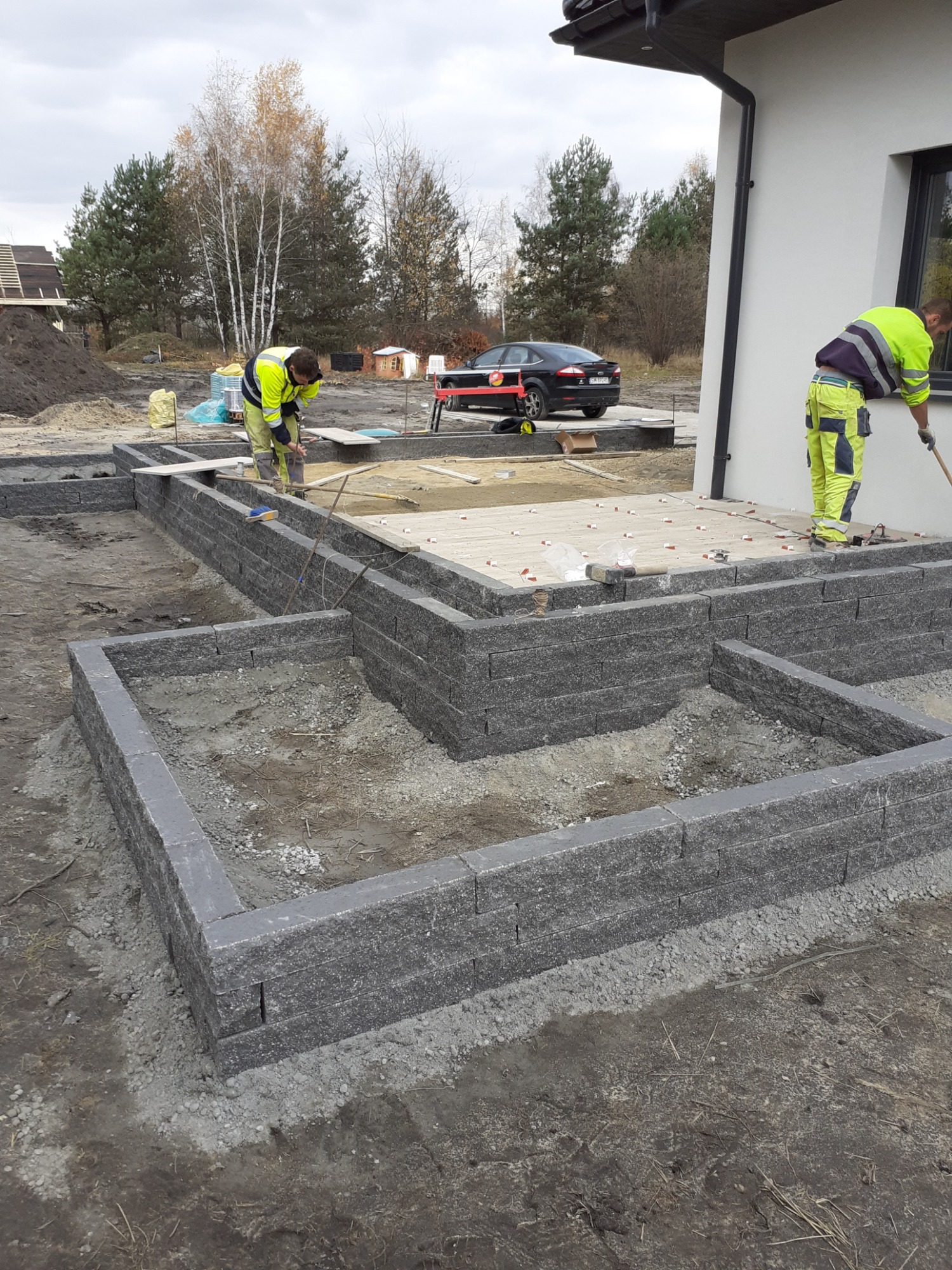 Budowa fundamentów z szarej kostki betonowej na posesji, dwaj pracownicy w odblaskowych ubraniach wyrównują teren, w tle samochód osobowy i fragment budynku.