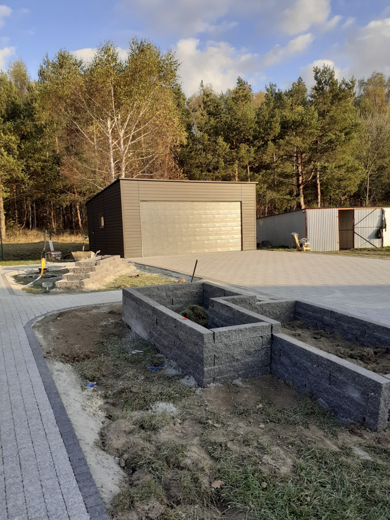 Układanie kostki brukowej na podjeździe z widocznym garażem blaszanym i konstrukcją pod rabaty kwiatowe, w tle drzewa.