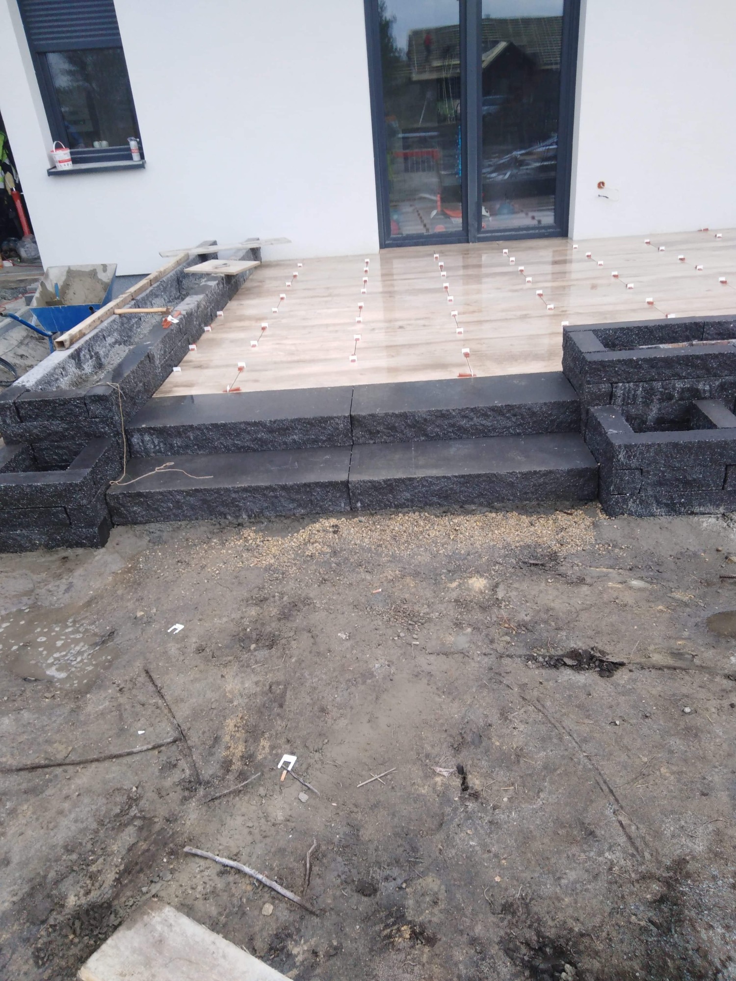 Budowa tarasu z drewnianą podłogą i schodami z ciemnego kamienia na zewnątrz budynku z białymi ścianami i dużymi oknami.