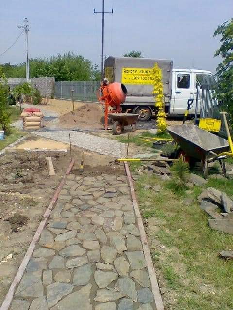 Układanie ścieżki z nieregularnych kamieni w ogrodzie, widoczny betonowy obrzeże, zaplecze budowy z betoniarką i workami cementu, trawa i rozrzucone kamienie.