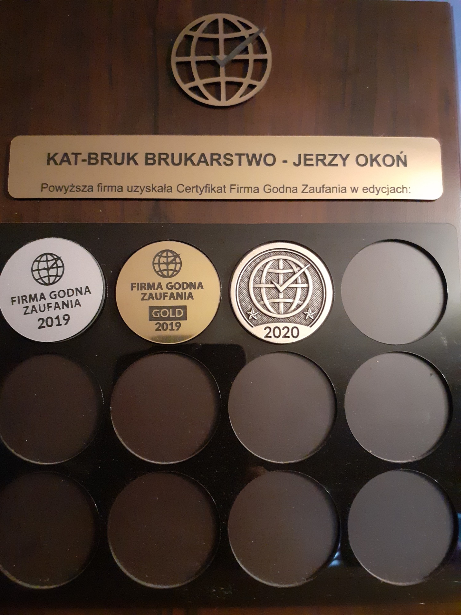 Tablica z certyfikatami 'Firma Godna Zaufania' dla firmy brukarskiej KAT-BRUK BRUKARSTWO - Jerzy Okoń, obejmująca lata 2019 (srebrny i złoty certyfikat) oraz 2020, umieszczona na ciemnobrązowym tle...