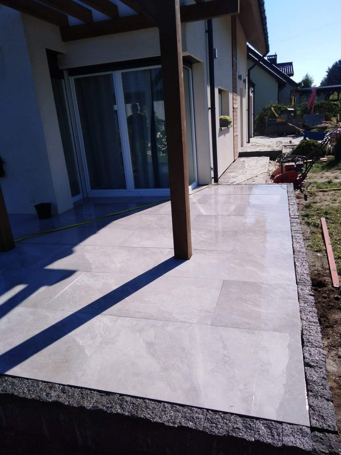 Wyłożony jasnoszarymi płytkami taras z drewnianą pergolą w tle, widoczny zagęszczarka wibracyjna i obrzeże z kostki granitowej.