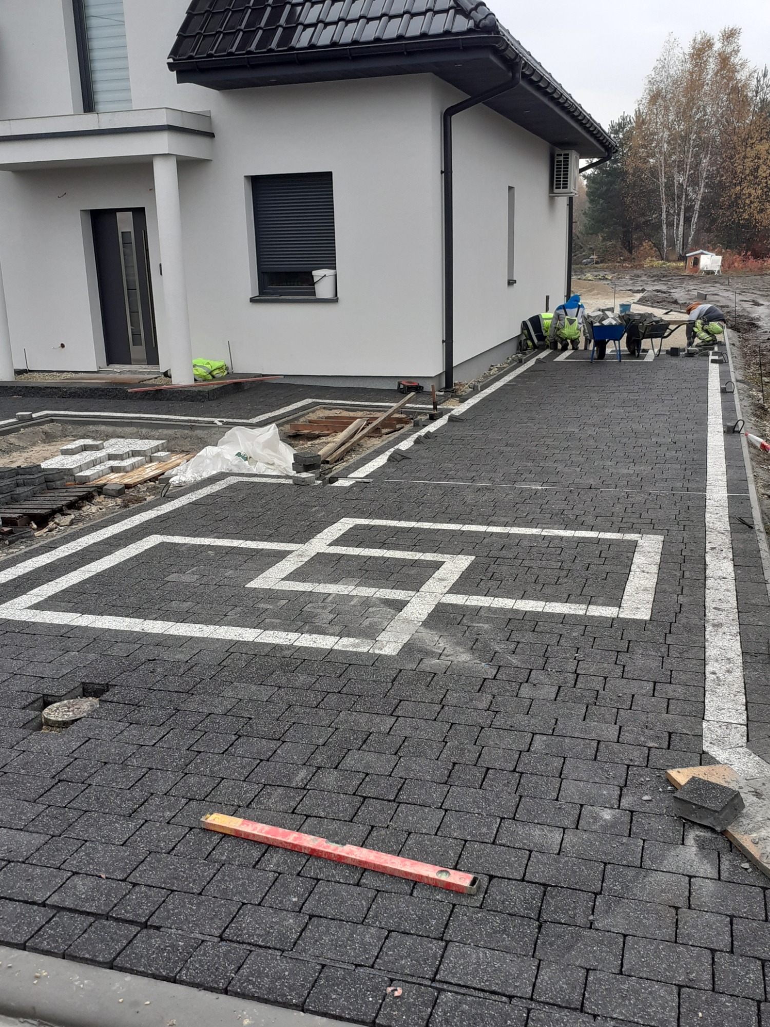 Układanie kostki brukowej w geometryczny wzór przed nowoczesnym domem; poziomica leży na nawierzchni, w tle pracownicy z taczkami.