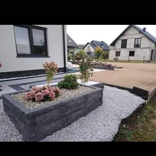 Ogródek z szarej kostki brukowej z kwiatami i kamieniami na tle domu i przygotowanego terenu pod budowę drogi dojazdowej.