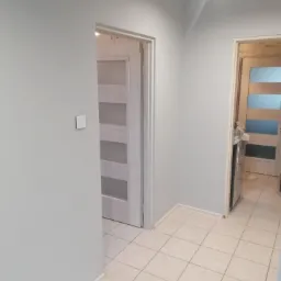 Jasnoszary korytarz po odświeżeniu, widoczne dwie pary drzwi w ościeżnicach, kremowe płytki podłogowe i biała listwa przypodłogowa