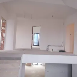 Wnętrze domu w trakcie prac wykończeniowych; widoczne surowe ściany pomalowane na biało, schody z wylewki betonowej, niezamaskowane otwory drzwiowe z fragmentami cegieł i narzędzia na podwyższeniu.