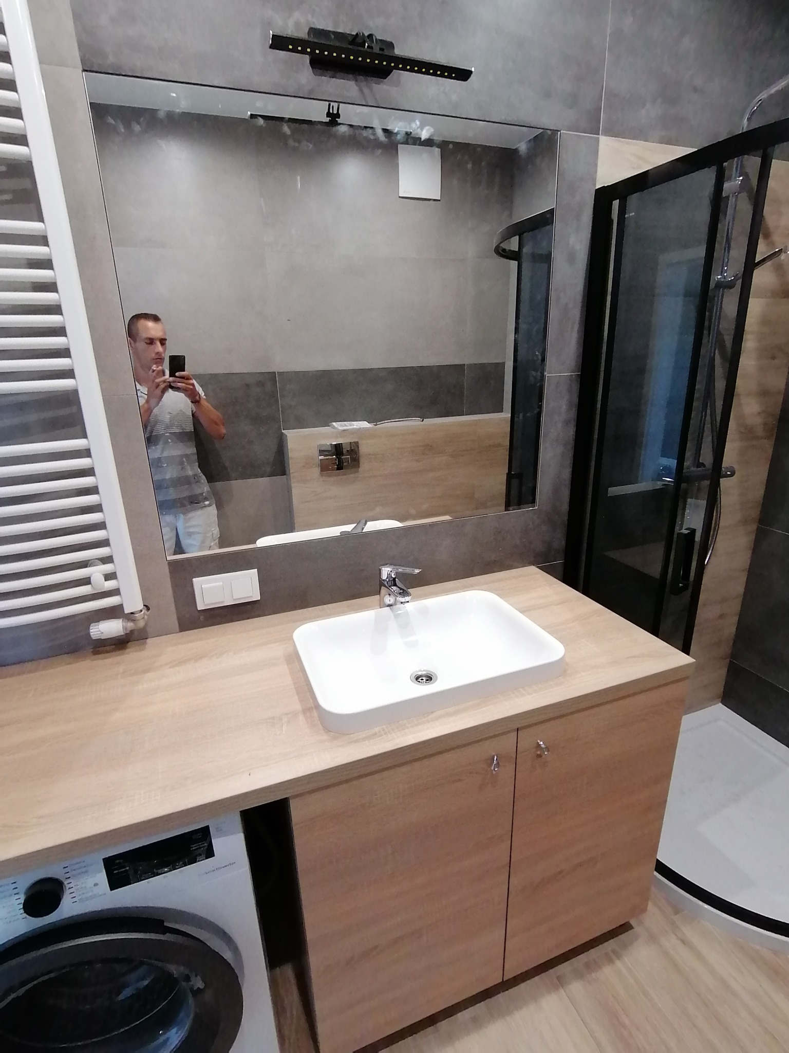 Nowoczesna łazienka z kabiną prysznicową, umywalką na szafce z blatem imitującym drewno oraz pralką w zabudowie; odbicie fotografa w lustrze.
