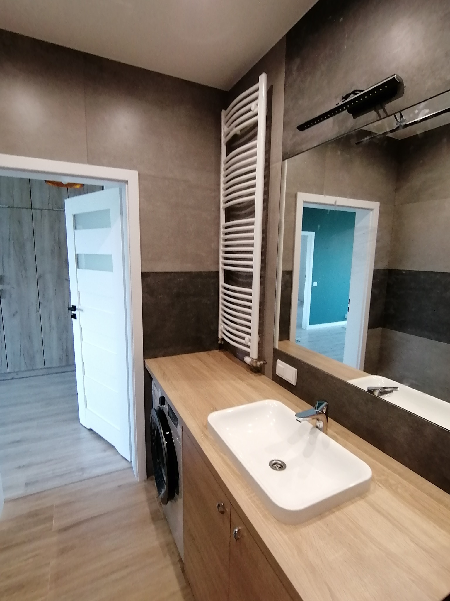 Nowoczesna łazienka z umywalką nablatową, grzejnikiem drabinkowym, pralką pod blatem i lustrem z oświetleniem, widok na otwarte drzwi do innego pomieszczenia z turkusową ścianą.