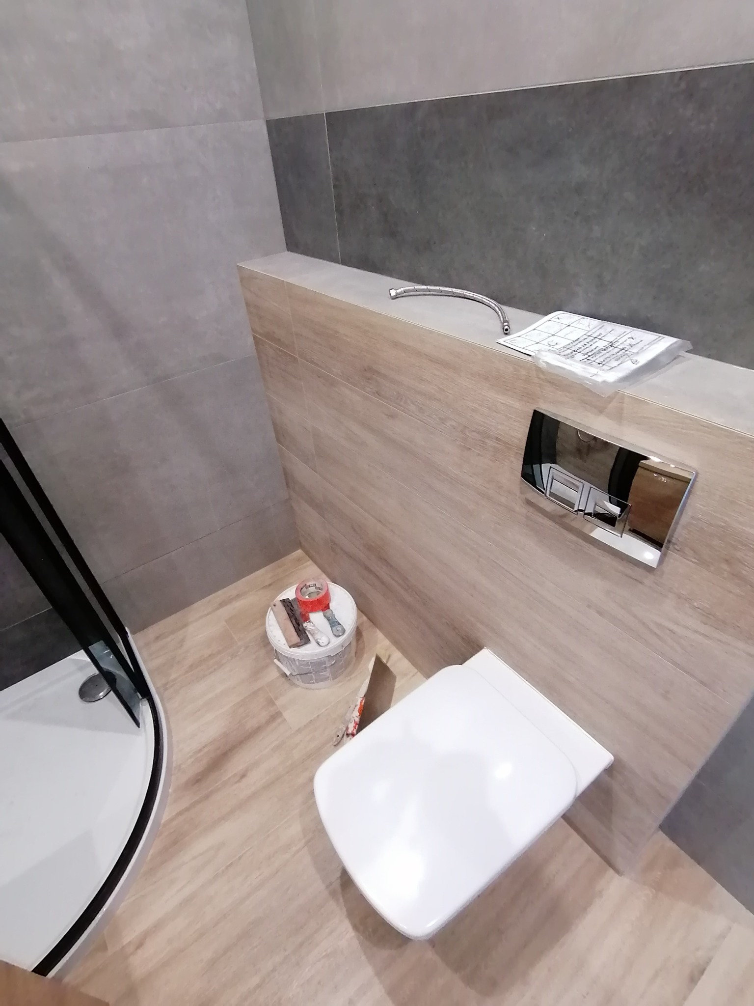 Wnętrze łazienki w trakcie montażu, widoczny stelaż podtynkowy z przyciskiem spłukującym, toaleta, fragment kabiny prysznicowej, wiadro z narzędziami i materiałami budowlanymi oraz wężyk dopływowy