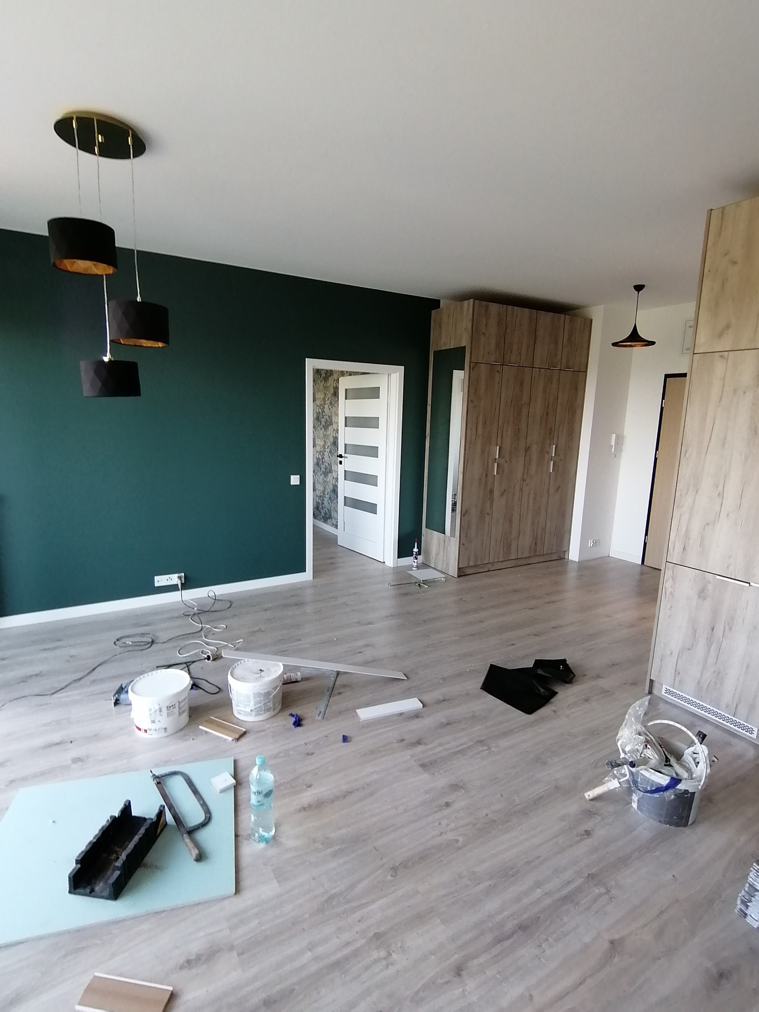 Wnętrze w trakcie prac wykończeniowych: pomalowana na ciemnozielono ściana, panele na podłodze, widoczne narzędzia i materiały budowlane, w tle szafa w zabudowie i drzwi.
