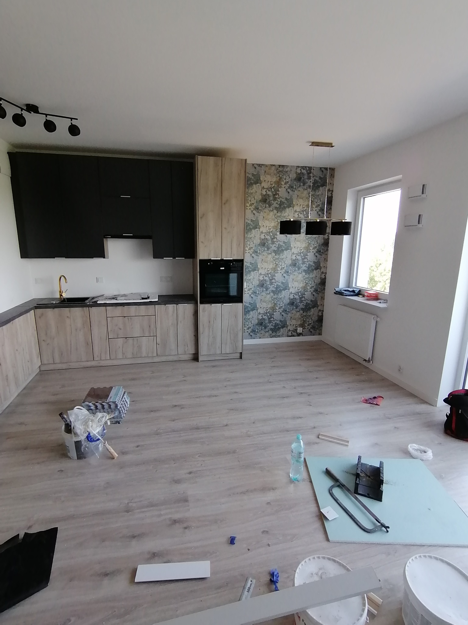 Wnętrze w trakcie prac wykończeniowych: widoczna zabudowa kuchenna z czarnymi szafkami górnymi i drewnopodobnymi dolnymi, fragment ściany z wzorzystą tapetą, narzędzia i materiały budowlane...