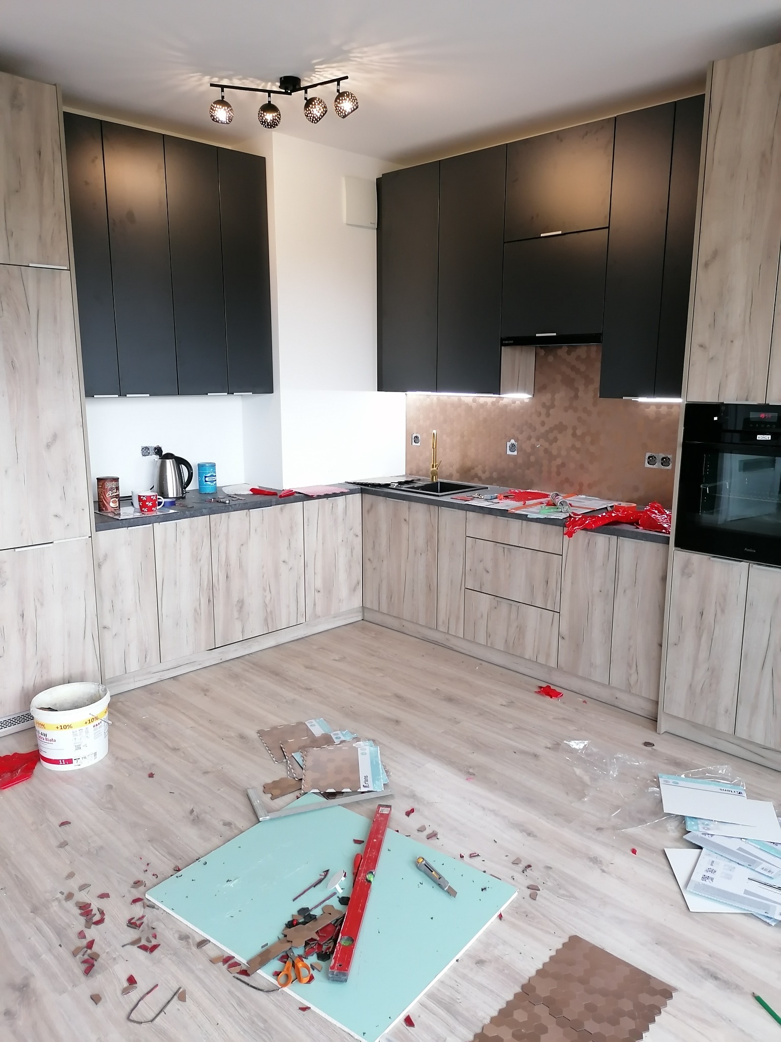 Montaż zabudowy kuchennej z drewnopodobnymi frontami i czarnymi szafkami wiszącymi, widoczne narzędzia i fragmenty mozaiki na podłodze, sugerujące prace wykończeniowe.