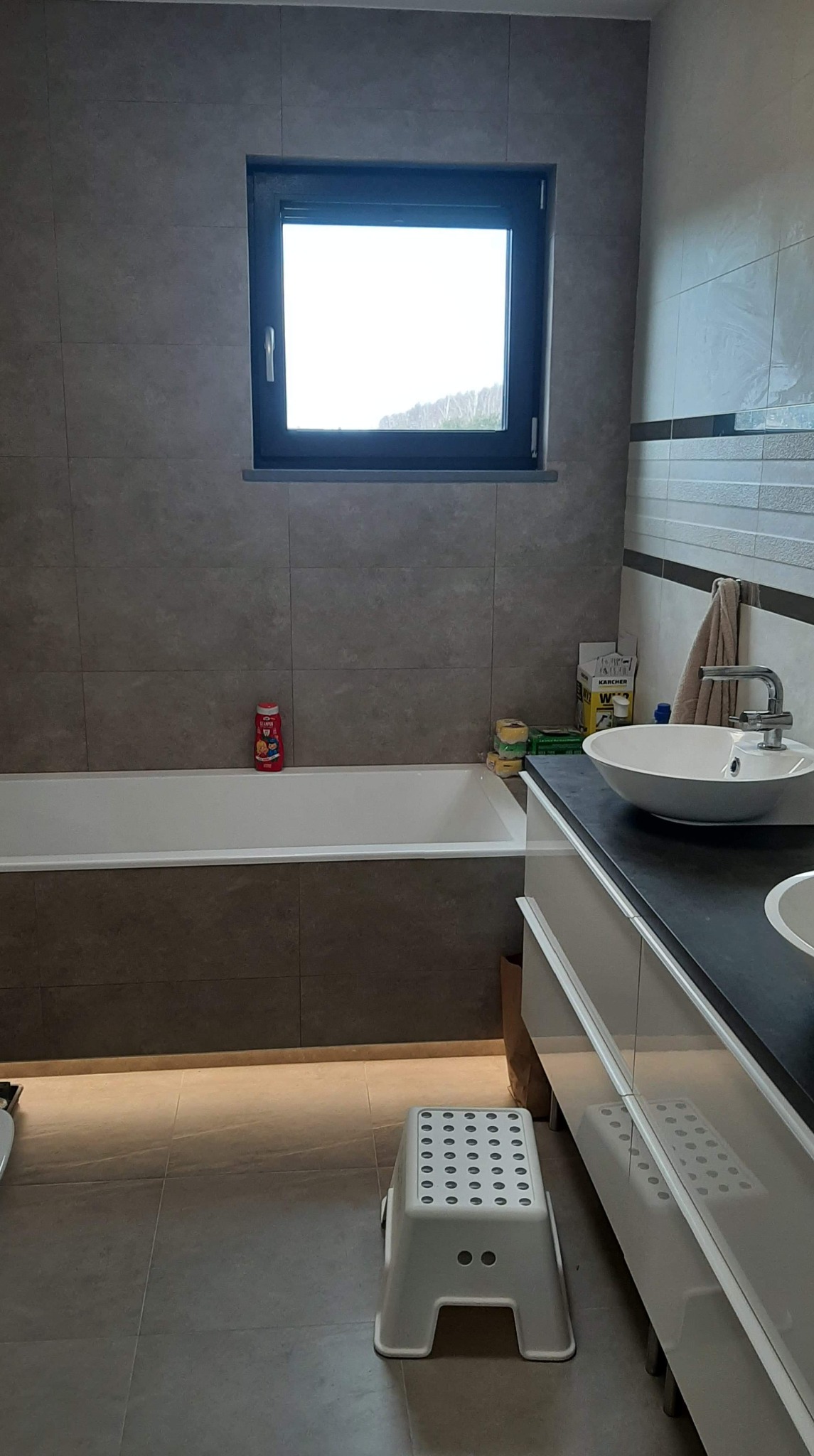 Nowoczesna łazienka z szarymi płytkami, białą wanną i umywalką nablatową, oświetleniem pod szafką i widokiem z okna na zalesione wzgórze.