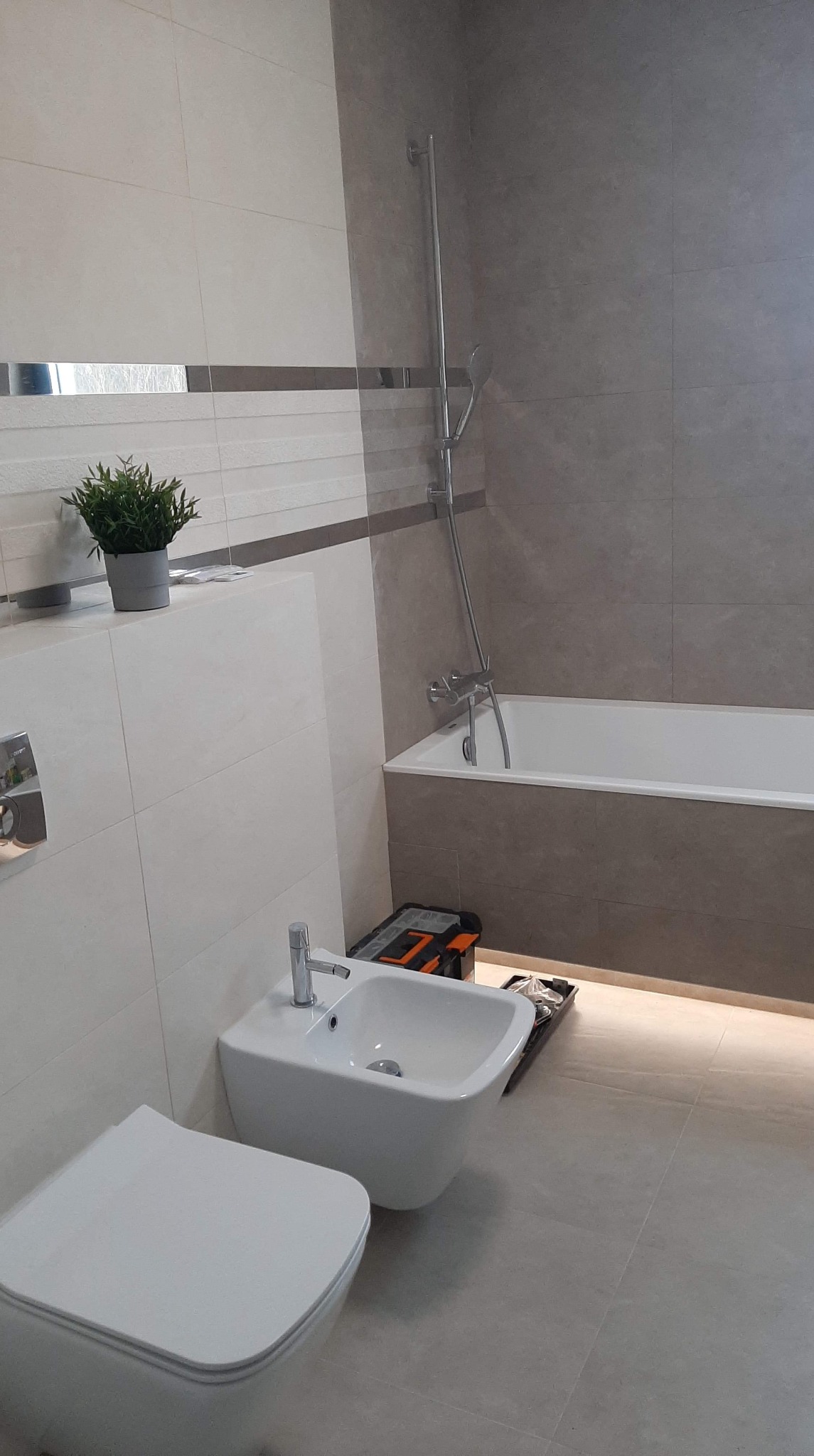 Nowoczesna łazienka w szaro-białych odcieniach z podświetleniem pod wanną, widoczna toaleta, bidet i skrzynka z narzędziami.