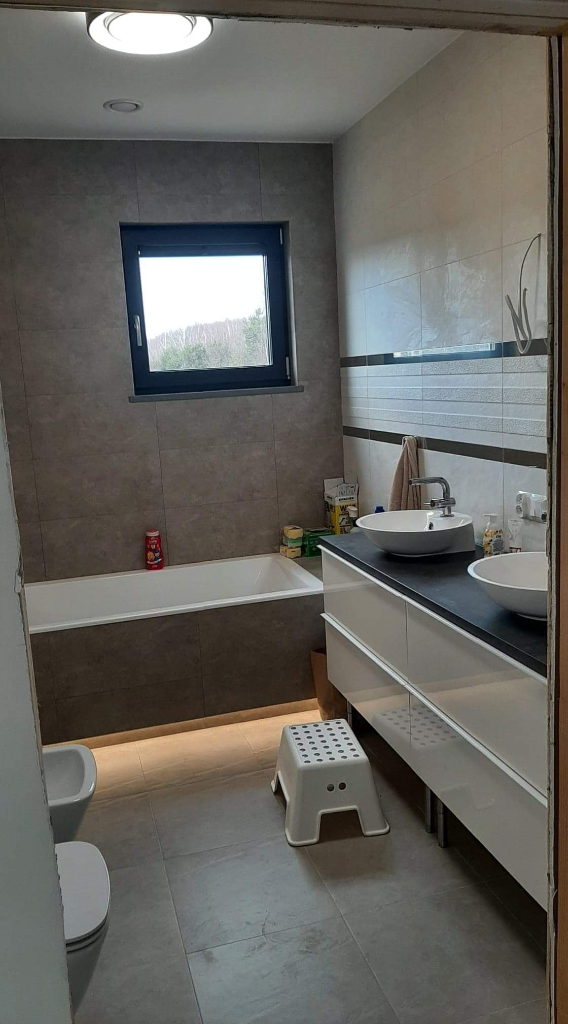 Nowoczesna łazienka z szarymi płytkami, podświetlaną wanną, białymi umywalkami nablatowymi i oknem z widokiem na drzewa, widziana z perspektywy wejścia.