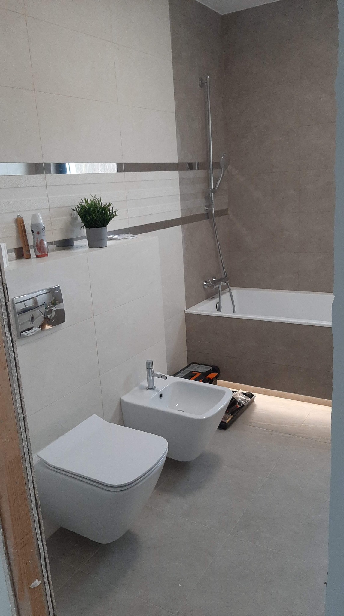 Nowoczesna łazienka w trakcie remontu, z podwieszaną toaletą i bidetem, widoczna skrzynka z narzędziami, płytki w odcieniach beżu i szarości, roślina doniczkowa na półce.