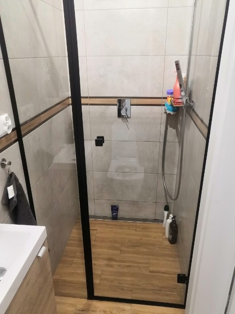 Nowoczesna kabina prysznicowa z czarną ramą, drewnopodobna podłoga, szare płytki z dekoracyjnym pasem, widoczne kosmetyki i akcesoria łazienkowe.