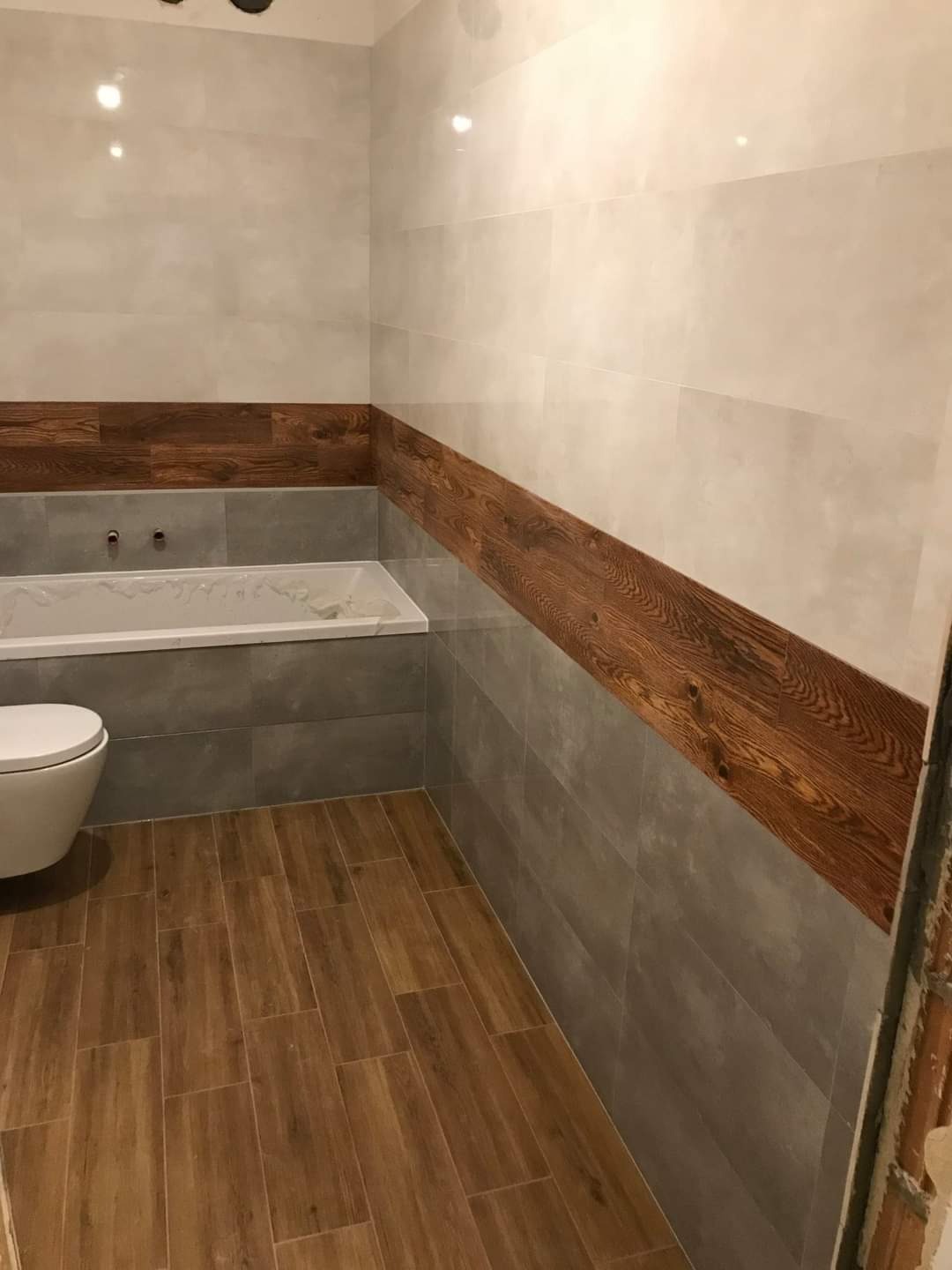 Wykończona łazienka z drewnopodobnymi panelami na podłodze i wzdłuż ściany, szare płytki do połowy wysokości, biała wanna i toaleta, widoczne niedokończone wykończenie przy drzwiach.