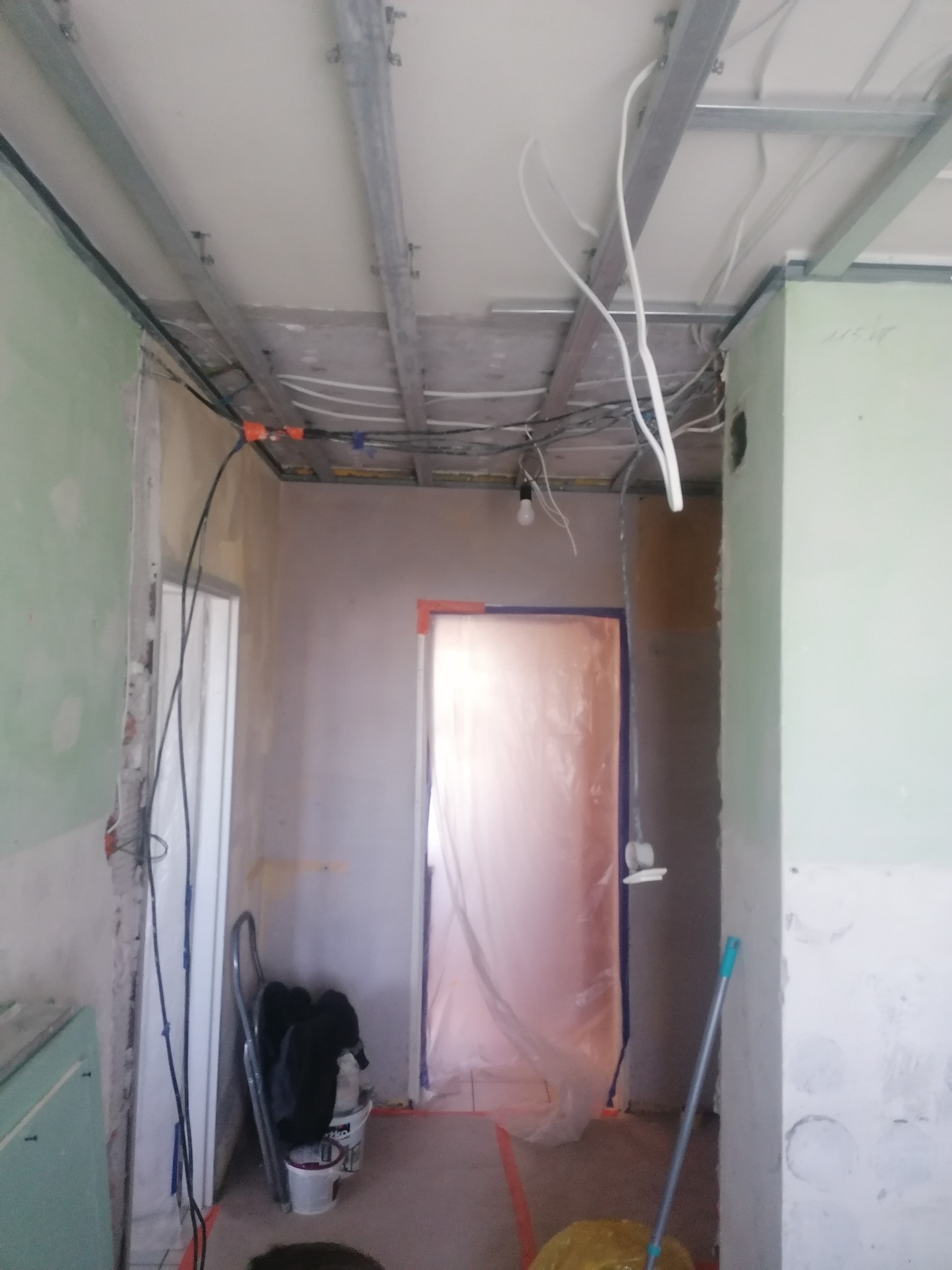 Wnętrze w trakcie prac: widoczne profile sufitowe, instalacje elektryczne, ściany przygotowane do malowania, zabezpieczone folią drzwi, drabina oparta o ścianę.