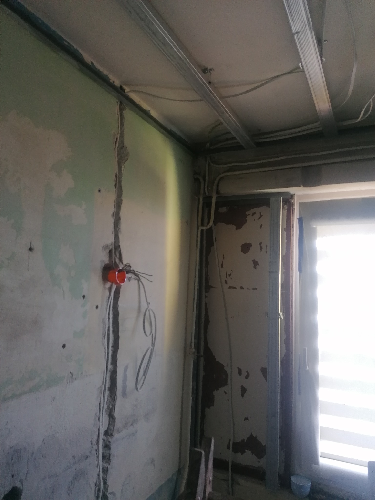 Ściana w trakcie remontu z widoczną instalacją elektryczną w bruzdach, profile pod zabudowę sufitu i fragment okna z roletą w tle.