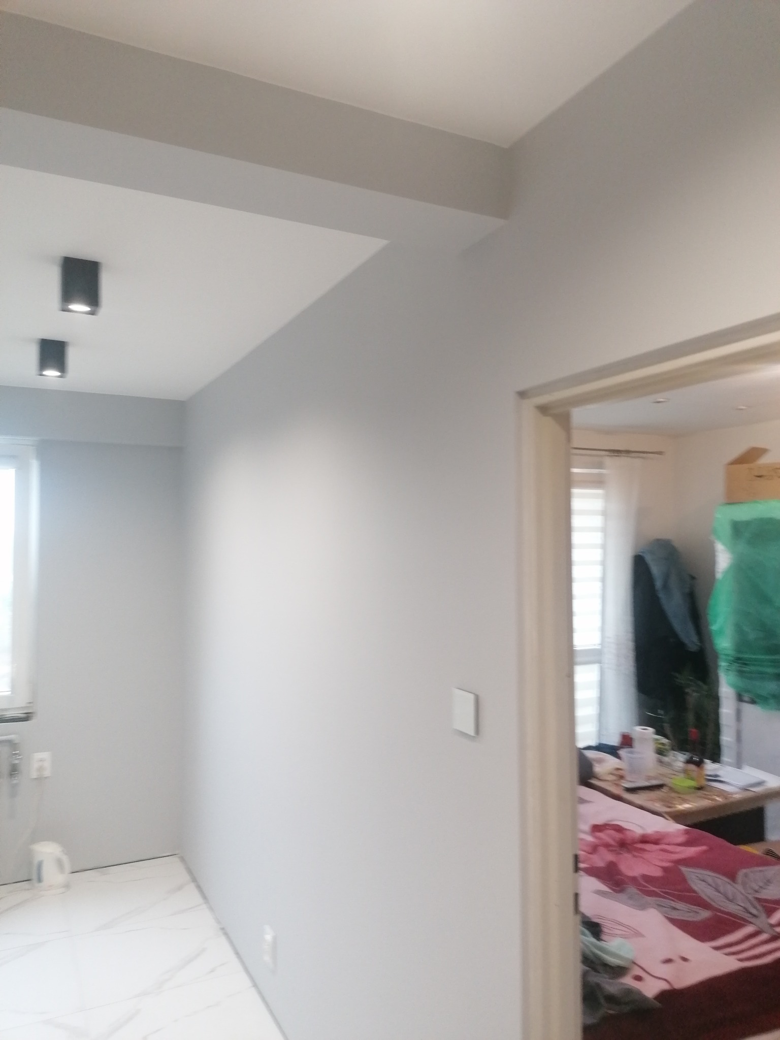 Wnętrze po malowaniu na szaro, widoczne białe marmurowe płytki podłogowe, czarne lampy sufitowe i fragment pokoju przez otwarte drzwi.