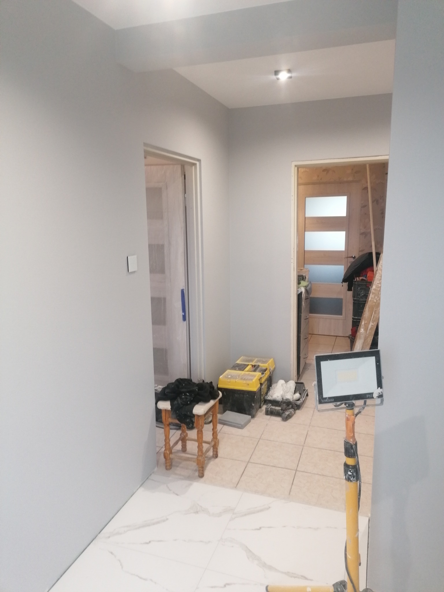 Korytarz w trakcie odświeżania: świeżo pomalowane szare ściany, białe płytki podłogowe z marmurkowym wzorem, widoczne narzędzia malarskie, lampa robocza STRUMM i krzesło z czarnym workiem...