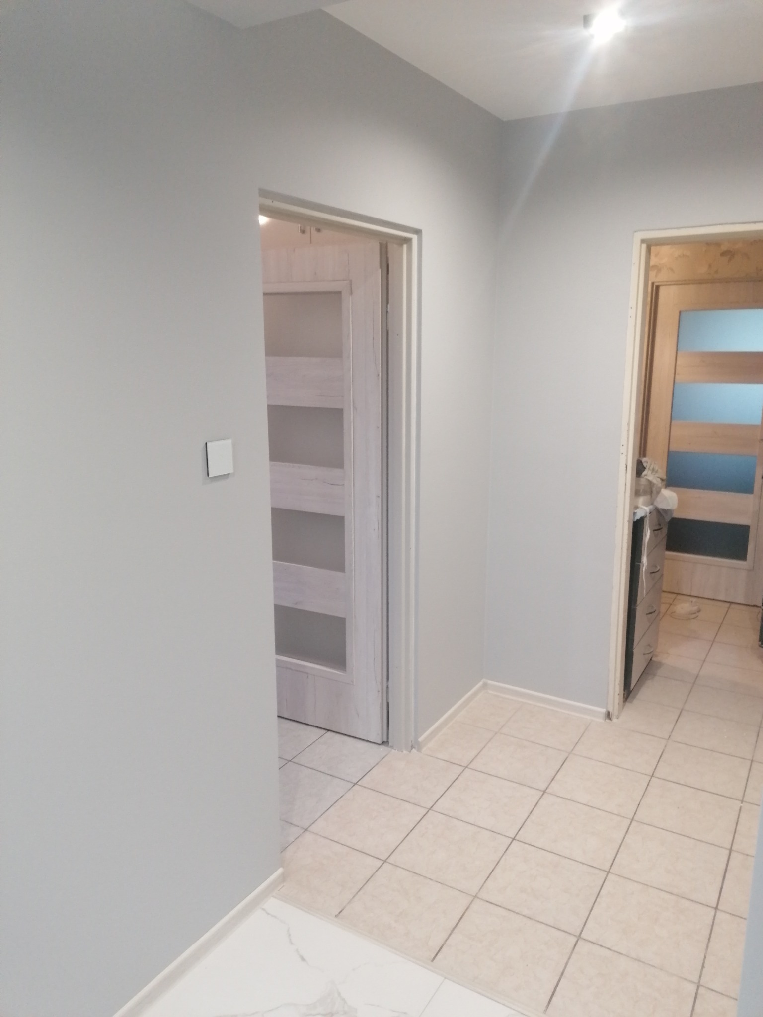 Jasnoszary korytarz po odświeżeniu, widoczne dwie pary drzwi w ościeżnicach, kremowe płytki podłogowe i biała listwa przypodłogowa