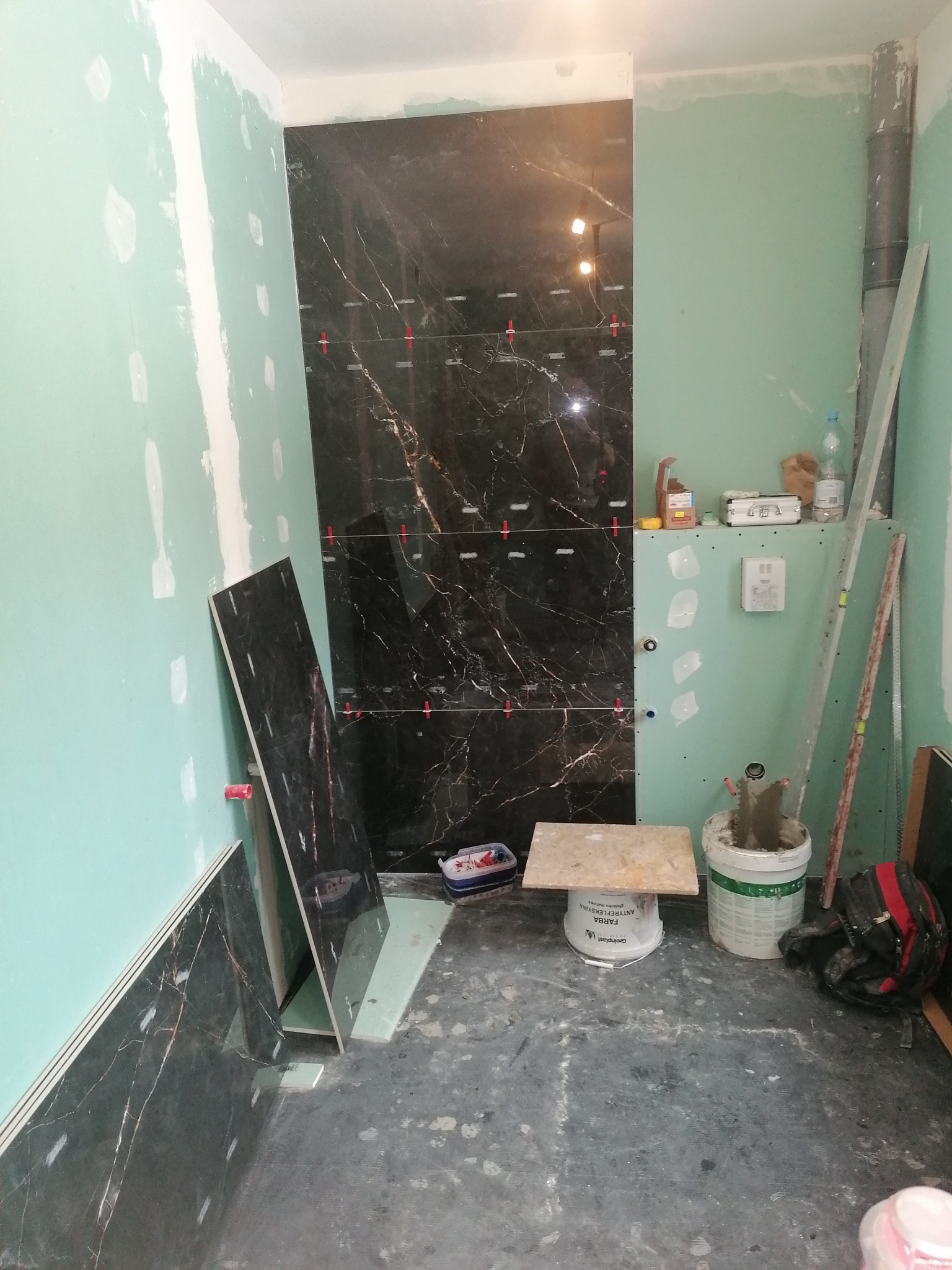 Łazienka w trakcie montażu płytek imitujących czarny marmur, z widocznymi krzyżykami dystansowymi i płytami gipsowo-kartonowymi na ścianach, narzędzia i materiały budowlane porozrzucane na podłodze.