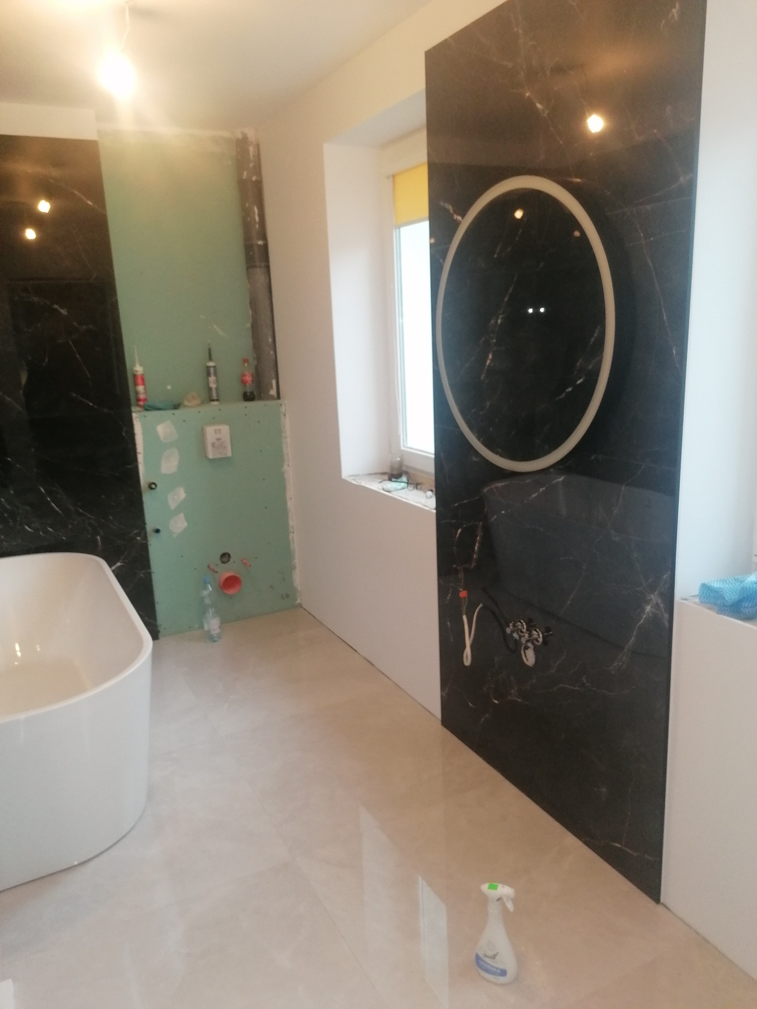 Wnętrze łazienki w trakcie prac wykończeniowych, widoczna wanna, ściana z czarnego marmuru z okrągłym lustrem z podświetleniem, oraz ściana z zielonej płyty gipsowej z widocznymi instalacjami...