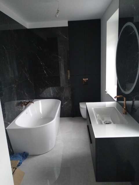 Nowoczesna łazienka z czarnymi marmurowymi ścianami, wolnostojącą białą wanną, umywalką z szafką w kolorze czarnym i okrągłym lustrem z podświetleniem LED.