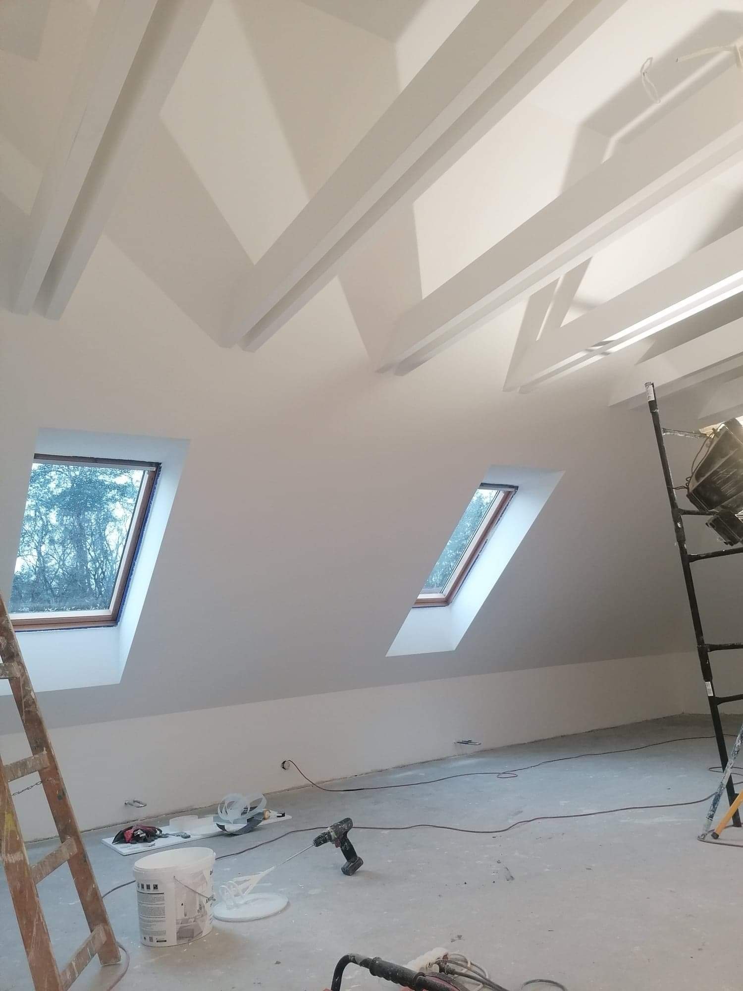 Poddasze po malowaniu na biało z widocznymi oknami dachowymi, drewnianymi belkami stropowymi i narzędziami malarskimi na podłodze.