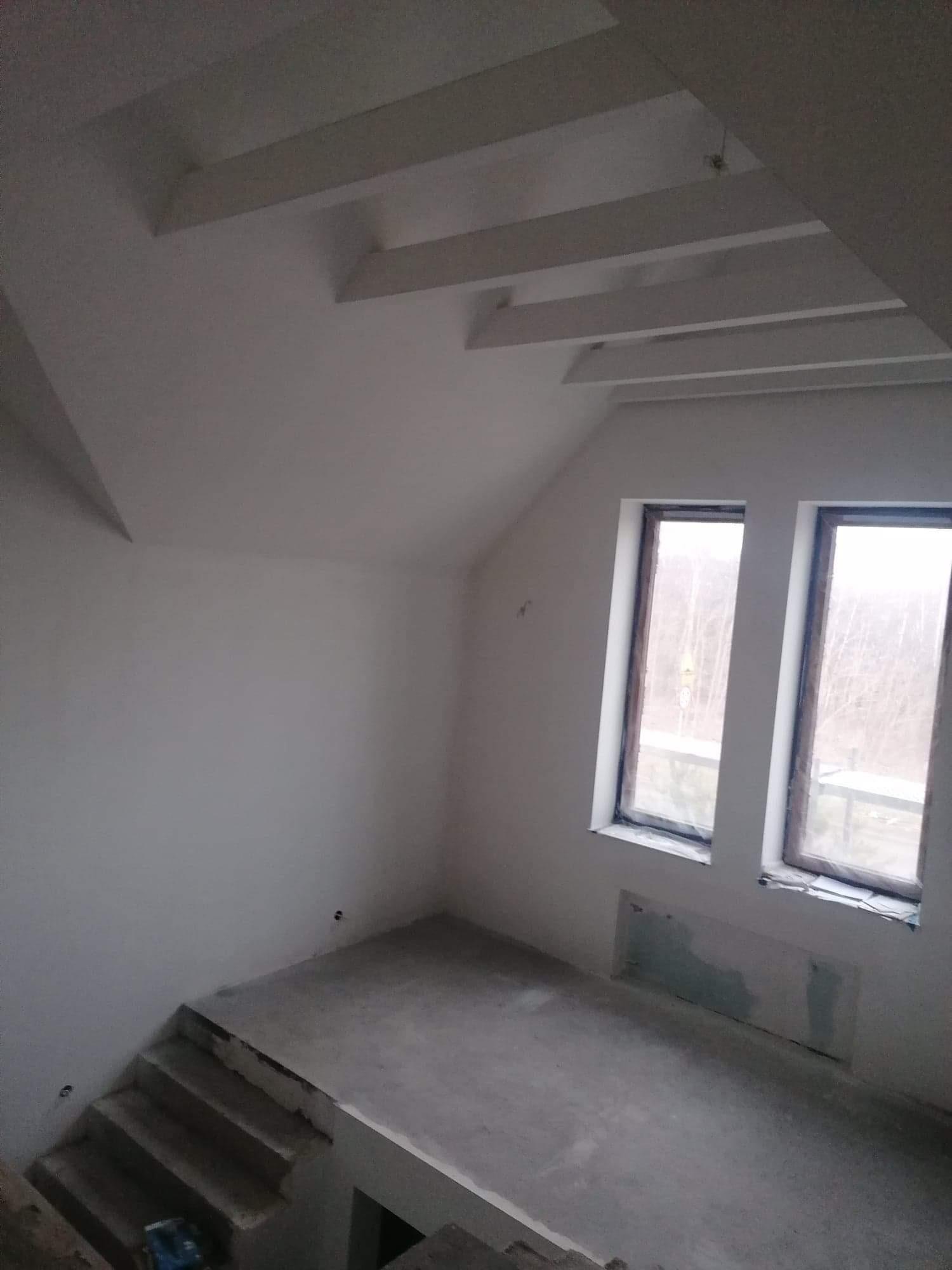 Surowe wnętrze poddasza z dwoma oknami, widocznymi schodami i przygotowaną podłogą, ściany i sufit pomalowane na biało, widoczne drewniane belki na suficie.