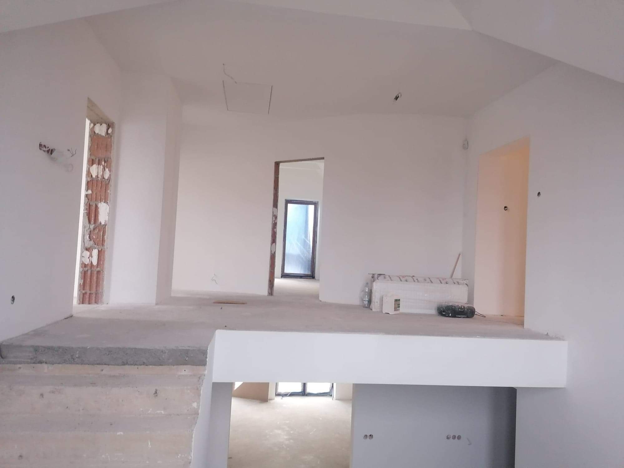 Wnętrze domu w trakcie prac wykończeniowych; widoczne surowe ściany pomalowane na biało, schody z wylewki betonowej, niezamaskowane otwory drzwiowe z fragmentami cegieł i narzędzia na podwyższeniu.