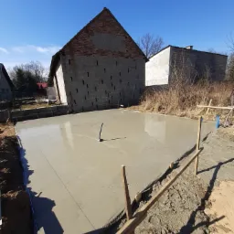 Wylany betonowy fundament pod budynek z wystającą rurą kanalizacyjną, drewniane szalunki wzdłuż krawędzi, w tle budynki gospodarcze i błękitne niebo.