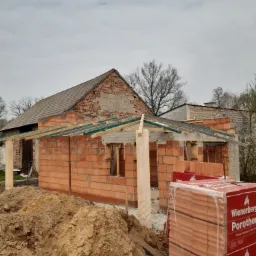 Budowa domu jednorodzinnego w trakcie murowania ścian z czerwonej cegły Porotherm, widoczne drewniane elementy konstrukcyjne dachu, w tle starszy budynek z cegły.