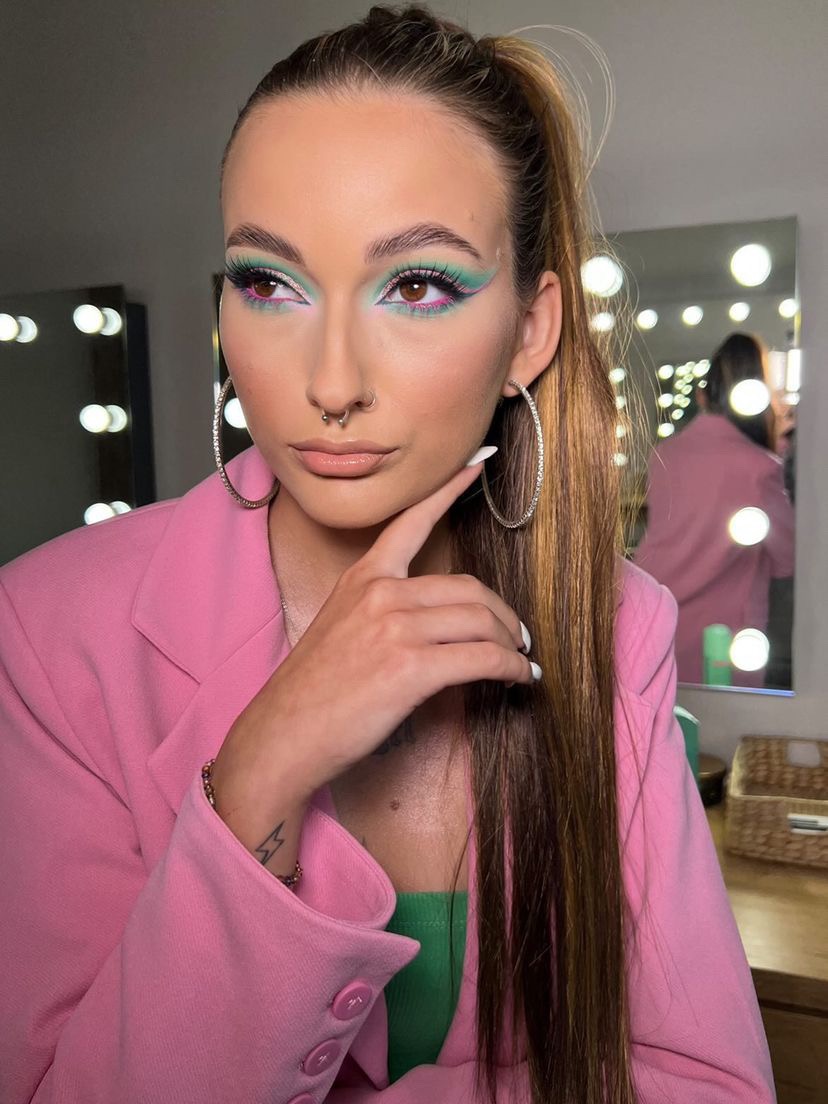 Portret kobiety z wyrazistym, kolorowym makijażem oczu w odcieniach różu, zieleni i błękitu, noszącej różowy żakiet i duże kolczyki koła, z włosami upiętymi w wysoki kucyk i piercingiem w nosie.