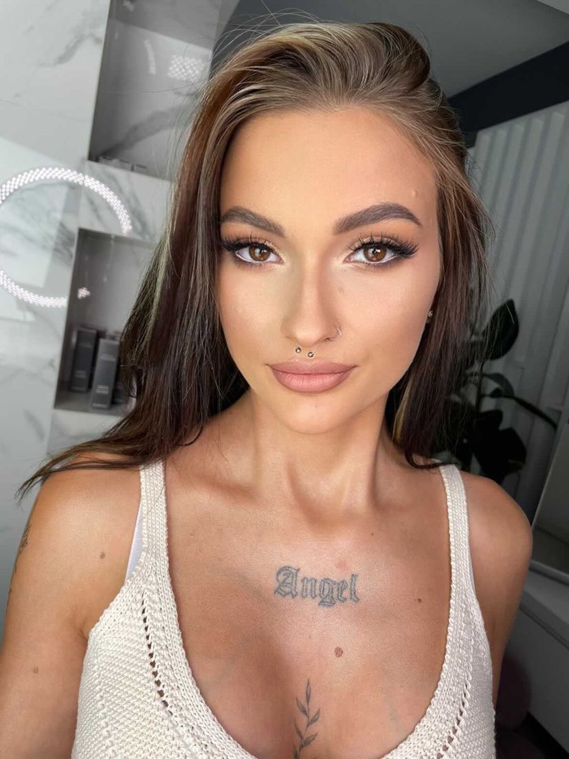 Portret kobiety z piercingiem w wardze, tatuażem 'Angel' na klatce piersiowej, w jasnej koszulce na tle marmurowej ściany i okrągłego lustra