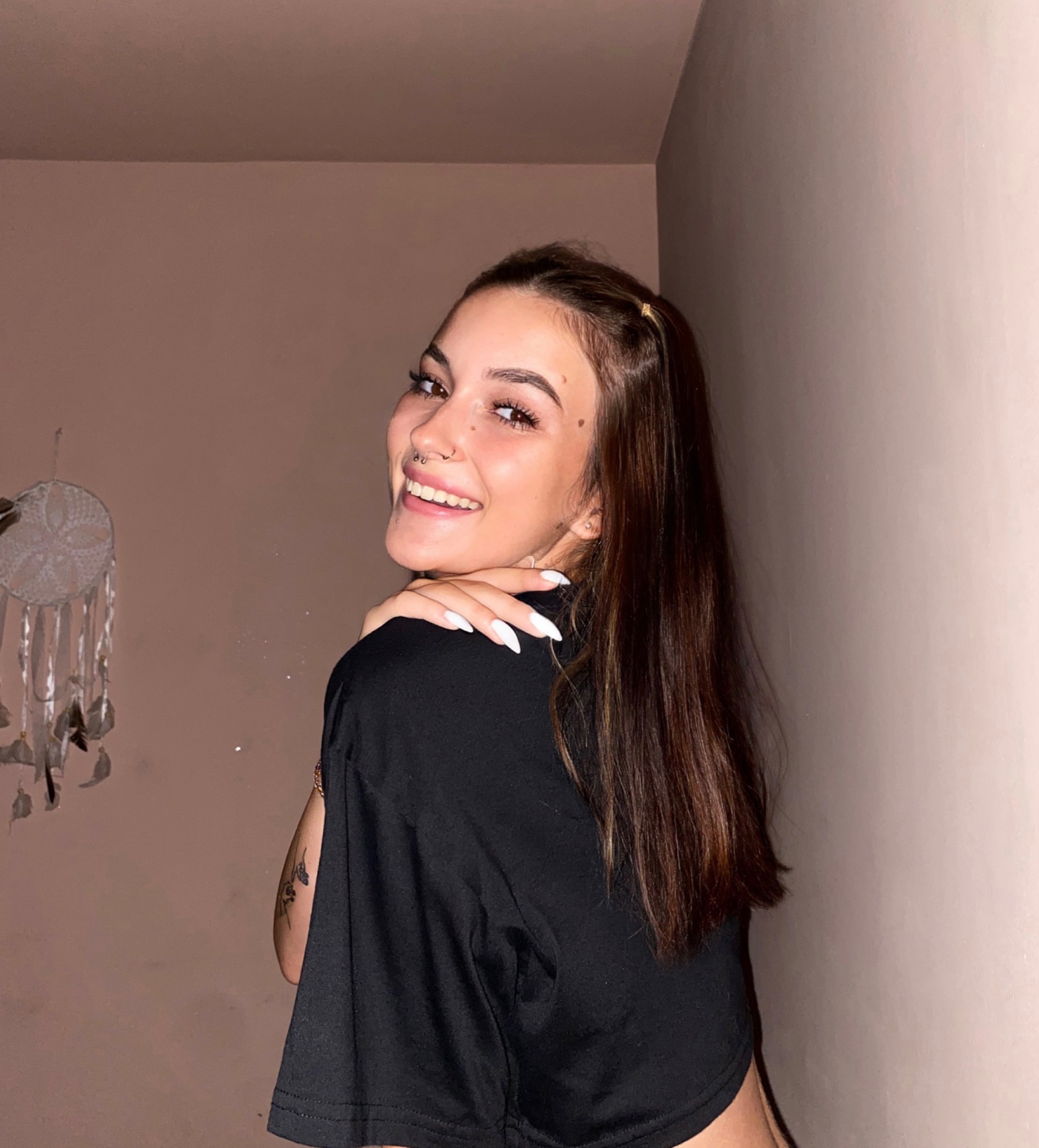 Uśmiechnięta młoda kobieta z piercingiem w nosie, spoglądająca przez ramię, z widocznym tatuażem na ramieniu i łapaczem snów w tle, w pomieszczeniu o ciepłym oświetleniu.