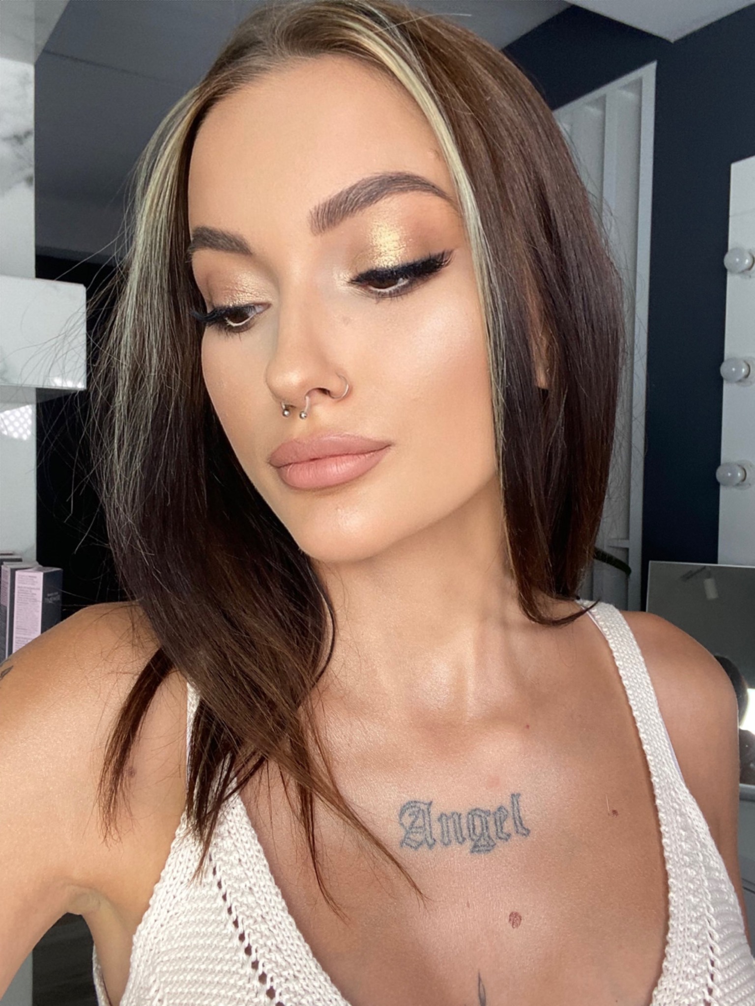 Portret kobiety z piercingiem w nosie, złotym makijażem oczu i tatuażem 'Angel' na dekolcie, patrzącej w dół.