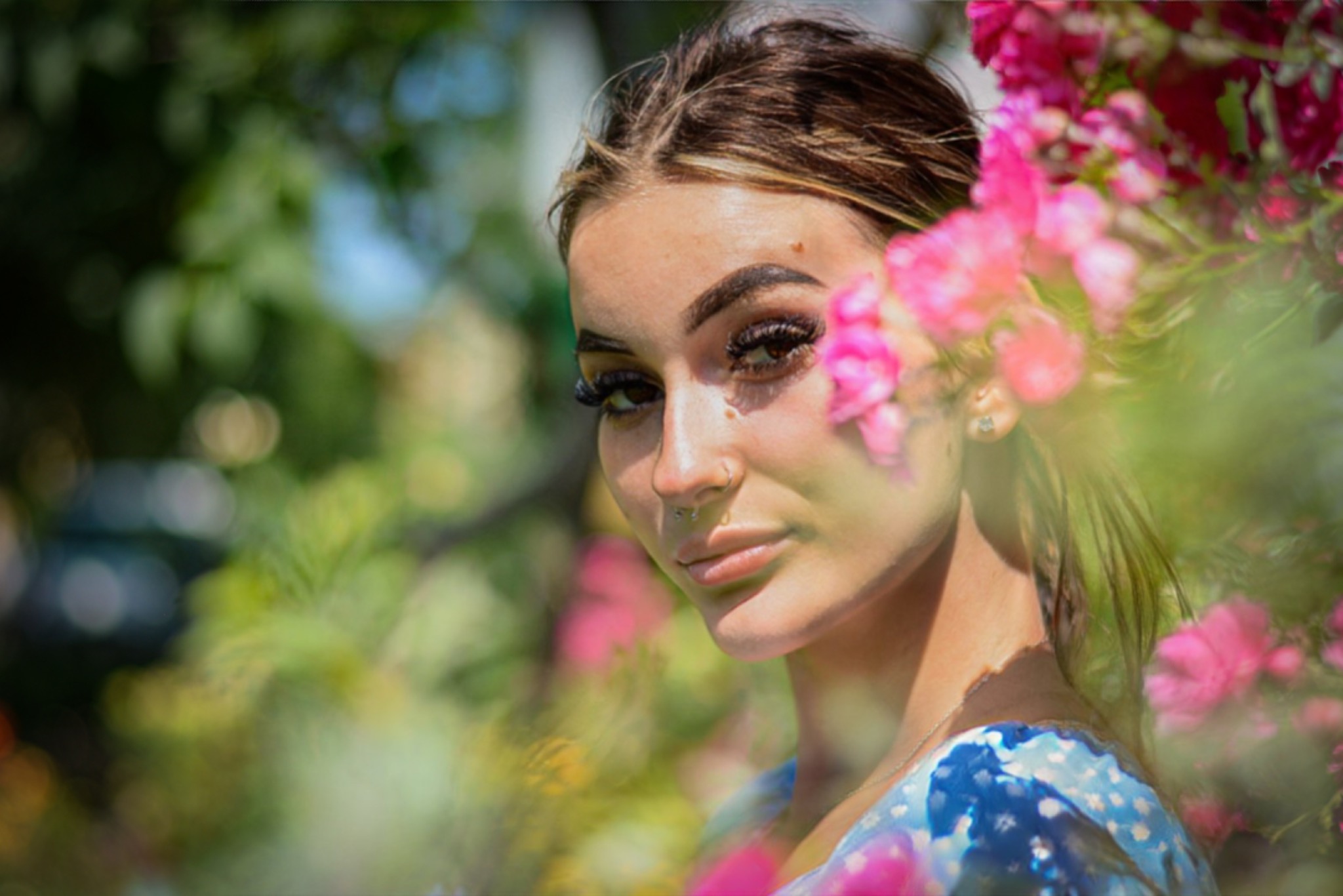 Portret młodej kobiety z piercingiem w nosie, otoczonej różowymi kwiatami i zielenią, w miękkim świetle.