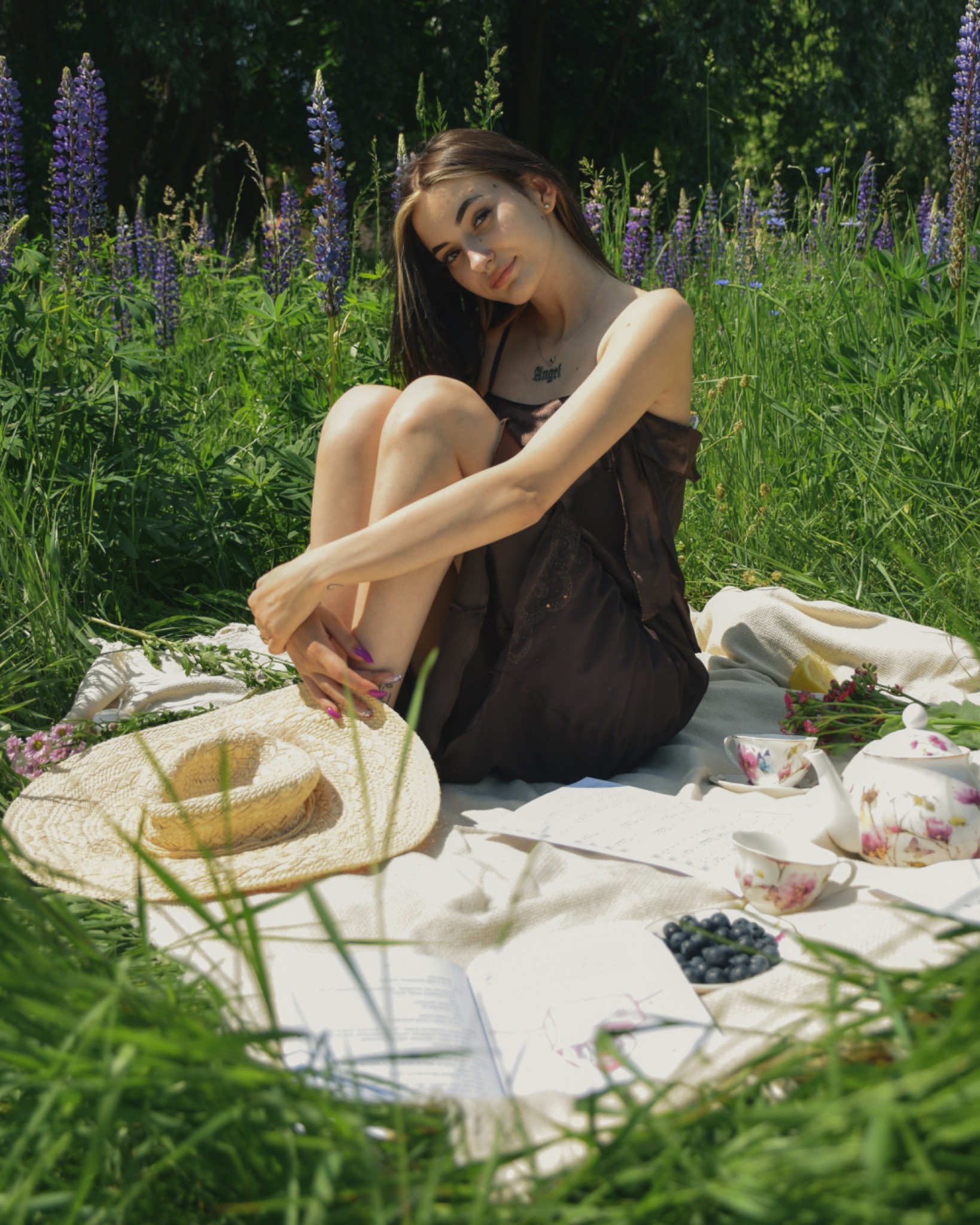 Młoda kobieta w brązowej sukience siedzi na kocu w trawie, otoczona kwiatami łubinu, z książką, kapeluszem, czajnikiem i filiżankami do herbaty oraz borówkami.