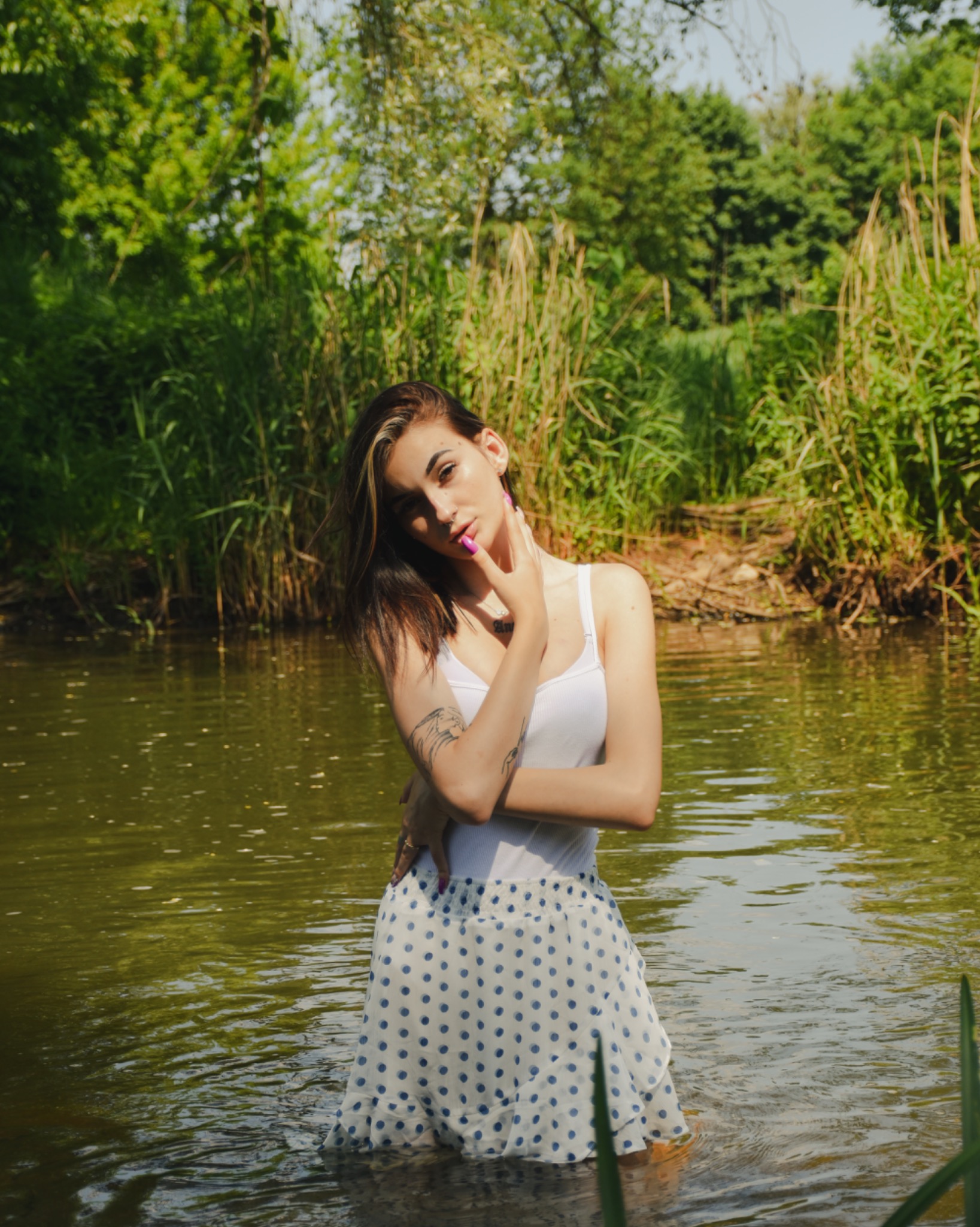 Kobieta w białym topie i spódnicy w kropki stoi w wodzie, z zaroślami i drzewami w tle, pozuje z ręką na szyi, eksponując tatuaż na ramieniu.
